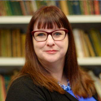A headshot of a woman in glasses smiling and looking into the camera with a blurred book shelf in the background